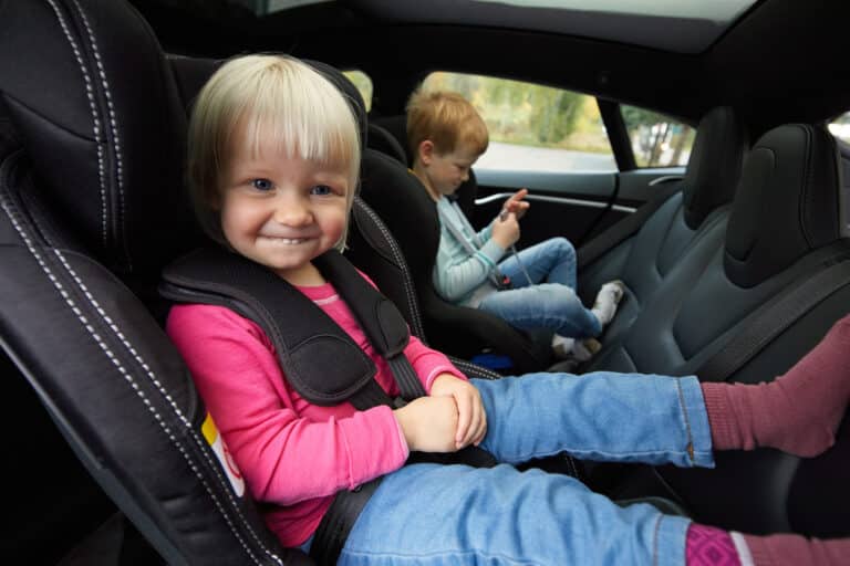 Jente sittende bakovervendt i bilstol ved siden av en gutt. Hun smiler.