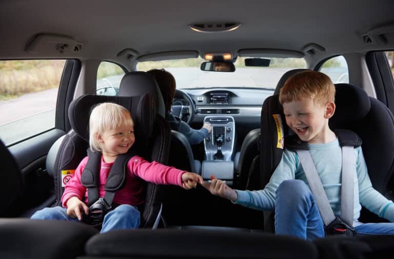 To barn sitter i bakovervendte bilstoler og fingrene møtes i midten av bilen.