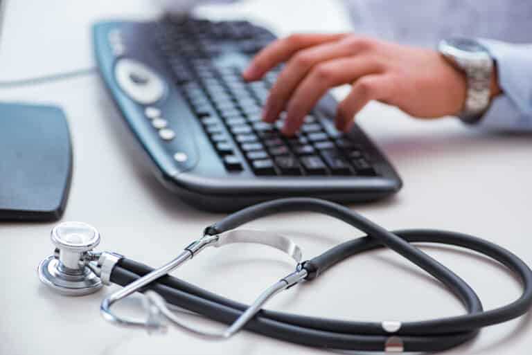 En doktor som sitter med en hånd på tastaturet. På siden av tastaturet ligger et stetoskop.