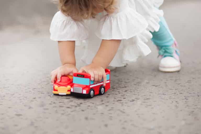 Liten jente som står bøyd mens hun holder seg fast i to lekebiler.