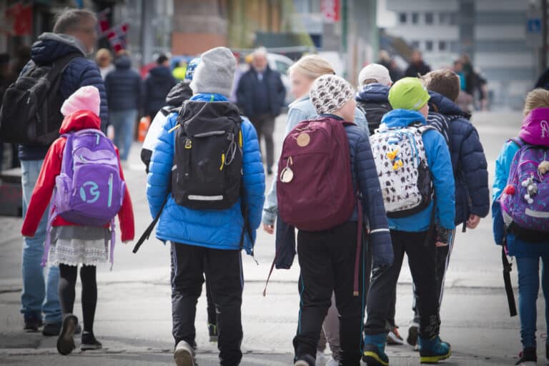 Mange skolebarn med ryggsekk på som går på en vei sammen med en voksen.