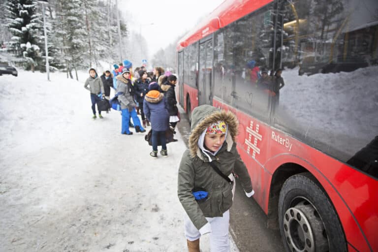 Barn står på en busstopp i snøvær, og en rød buss venter.