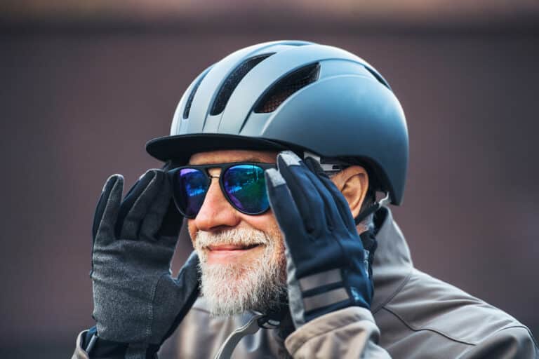 Mann med grått skjegg med solbriller, sykkelhjelm og hansker på. Han holder seg til brillene og er blid.