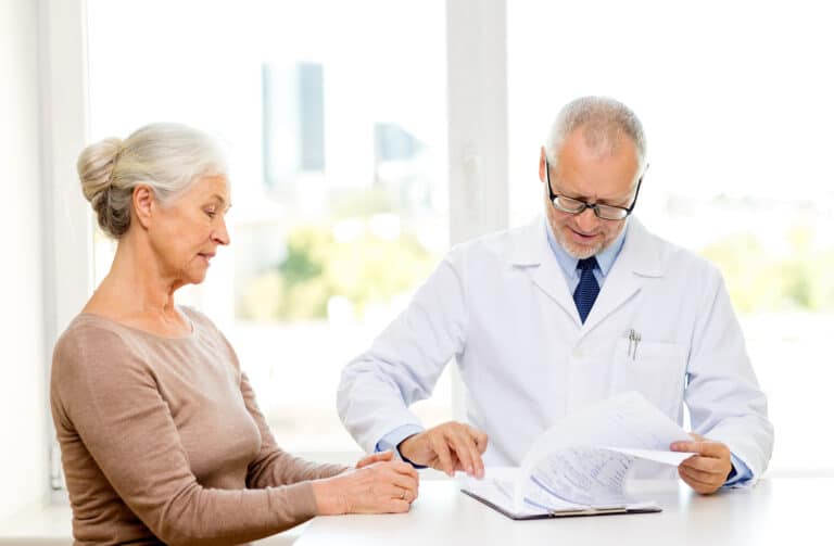 En eldre kvinne som sitter sammen med en lege. Legen blar i en legejournal og de prater sammen.