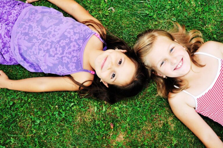 To jenter som ligger i gresset. De ser opp i linsa på kamera og smiler.