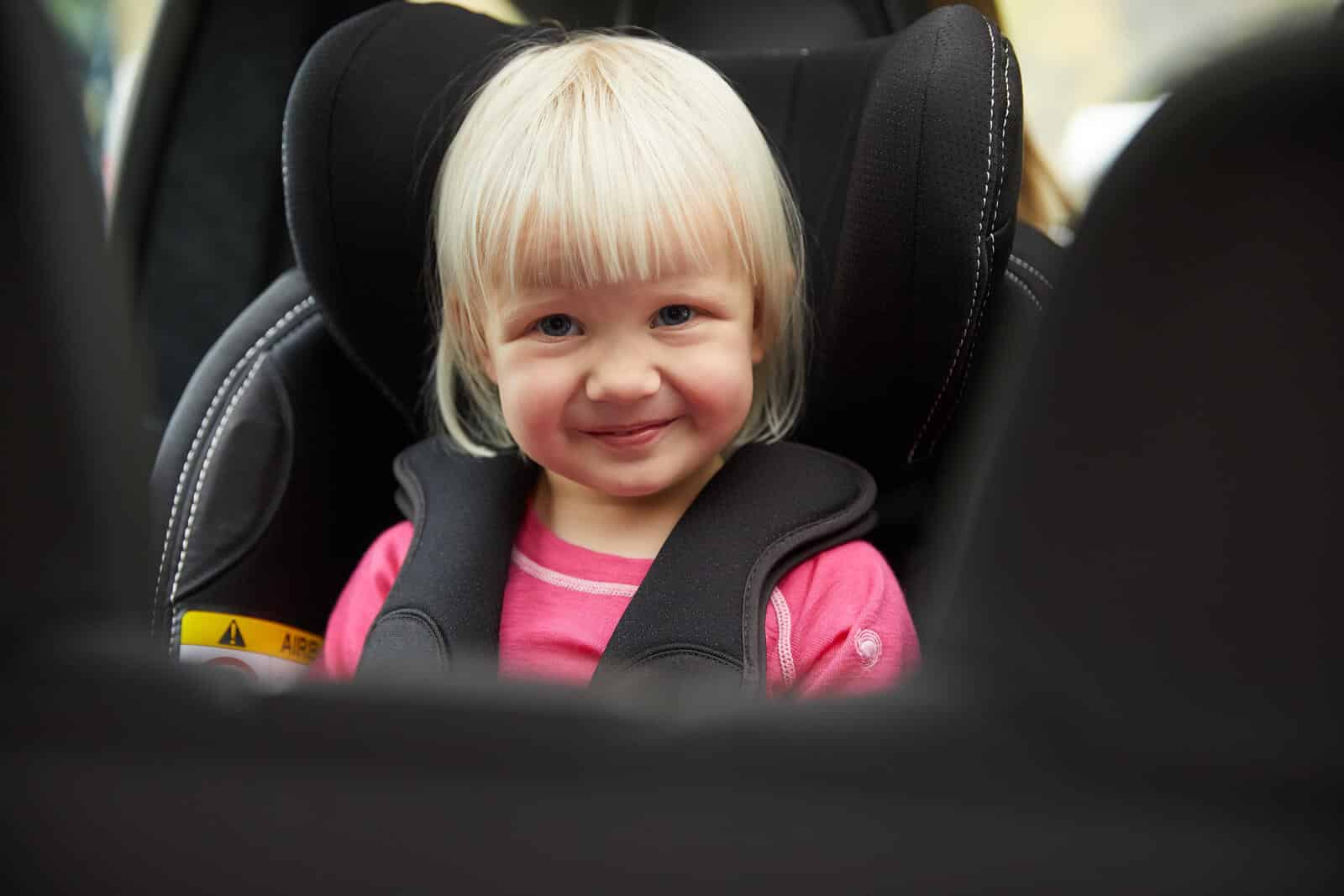Et barn sitter i bakovervendt bilstol og smiler mot kamera, som er i bagasjerommet.