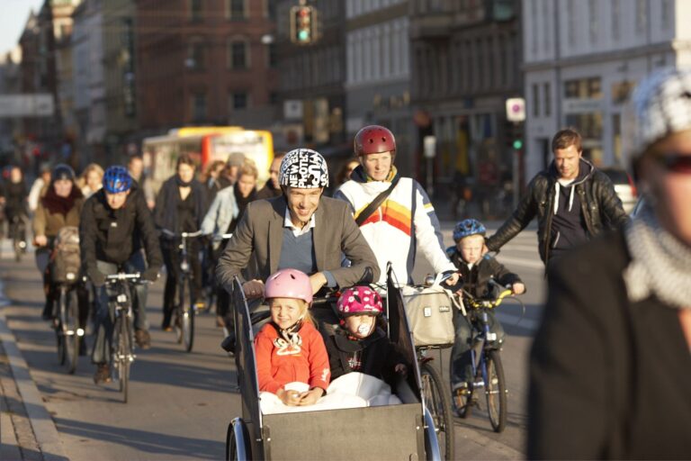 En transportsykkel i midten av en mengde syklister. Transportsykkelen har to barn i transportkassen og han som sykler dem smiler.