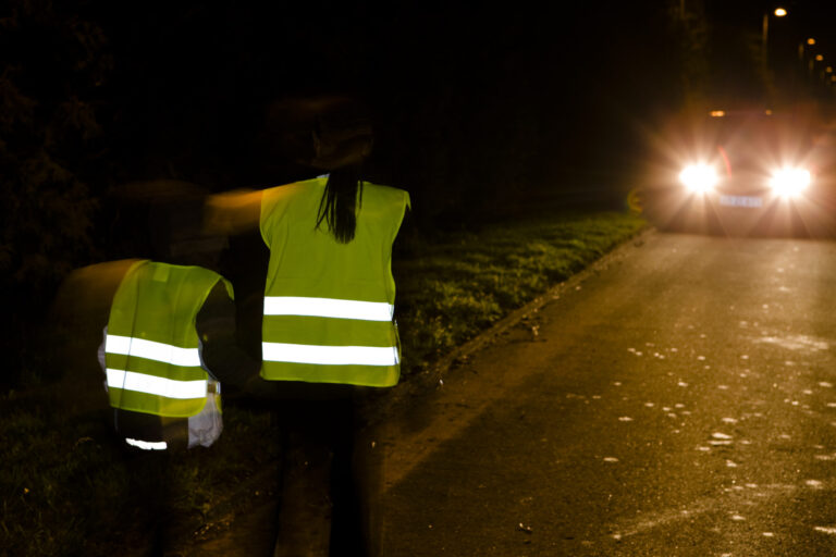 To personer med refleksvest som går på venstre side av veien i møte med en bil.